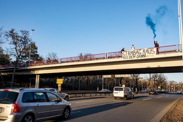 Drei Aktivist:innen stehen auf einer Brücke mit einem Transparent auf dem "Strabag raus aus dem Danni" steht, ein Aktivist hält eine blaue Nebelkerze