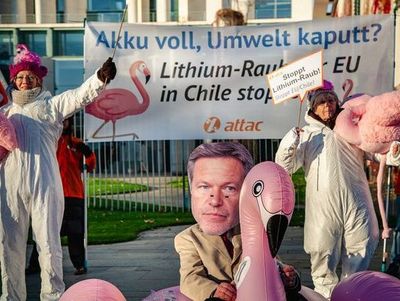Banner "Akku voll, Umwelt kaputt? Lithiumraub der EU in Chile stoppen! mit attac Logo und zwei Flamingos"; davor zwei Attacies in weißen Maleranzügen halten Plüschflamingos als Maskottchen der Kampagne, ein Attacie mit Habeck-Maske sitzt auf aufblasbarem Flamingo
