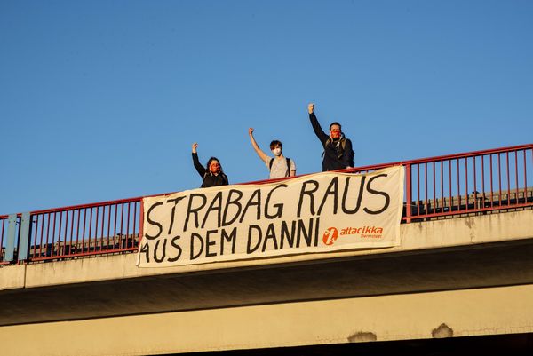 Drei Aktivist:innen stehen auf einer Brücke mit einem Transparent auf dem "Strabag raus aus dem Danni" steht