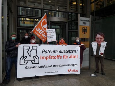 Attacies halten Banner "Patente aussetzen: Impfstoff für alle! Globale Solidarität statt Konzernprofite"; ein Aktivist mit Lauterbach-Maske hält ein zerrissenes "Patent" in den H#nden.